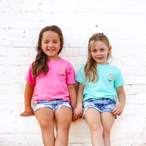 May include: Two young girls wearing matching t-shirts with a colorful circle design. The girl on the left is wearing a pink shirt and the girl on the right is wearing a light blue shirt. They are both wearing denim shorts and smiling at the camera.