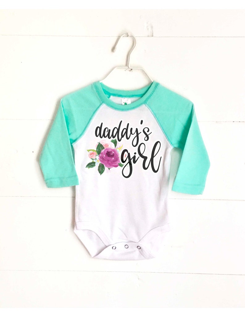 May include: White long-sleeved baby onesie with a mint green raglan sleeve. The onesie has a floral design and the text "daddy's girl" printed on the front.