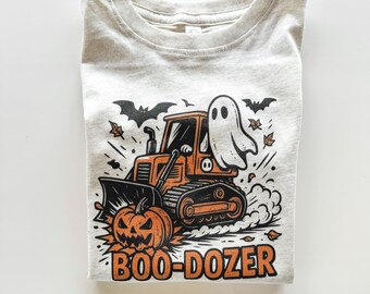 Peut inclure: Un t-shirt gris clair avec un motif sur le thème d'Halloween. Le motif comprend un bulldozer orange, une citrouille-lanterne, un fantôme et les mots "BOO-DOZER" en orange. Des chauves-souris et des feuilles noires complètent le design festif.