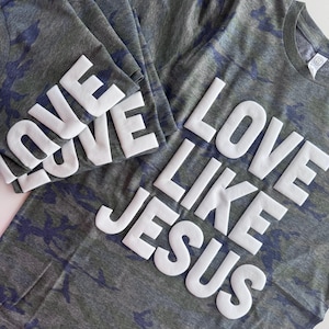 May include: Two short-sleeved t-shirts in a dark gray and blue camouflage pattern. The shirts feature the white, raised, block-letter text "LOVE LIKE JESUS" printed across the front. The fabric has a heathered texture and a crew neckline.