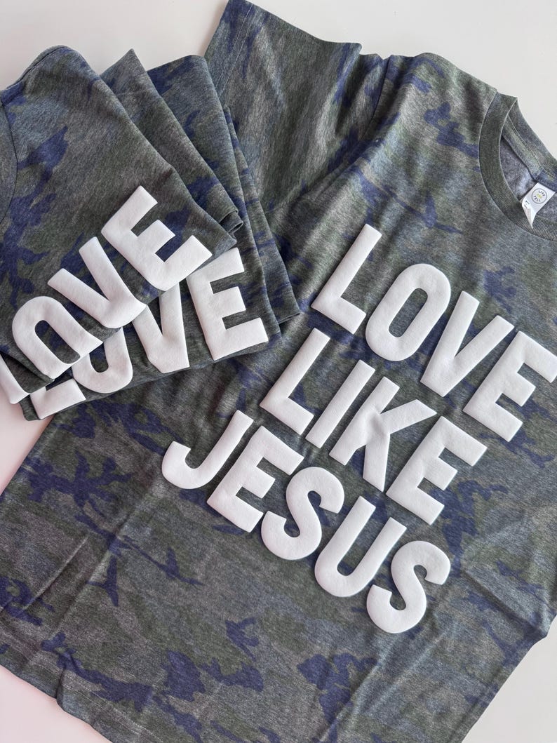 May include: Two short-sleeved t-shirts in a dark gray and blue camouflage pattern. The shirts feature the white, raised, block-letter text "LOVE LIKE JESUS" printed across the front. The fabric has a heathered texture and a crew neckline.