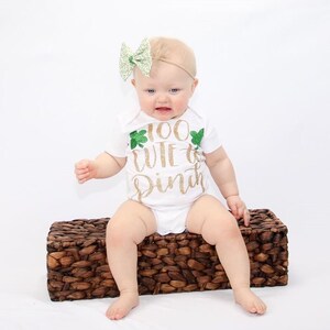 May include: A white baby onesie with a gold glitter design that says "Too Cute to Pinch" with green shamrock accents. The baby is wearing a green and white bow headband.