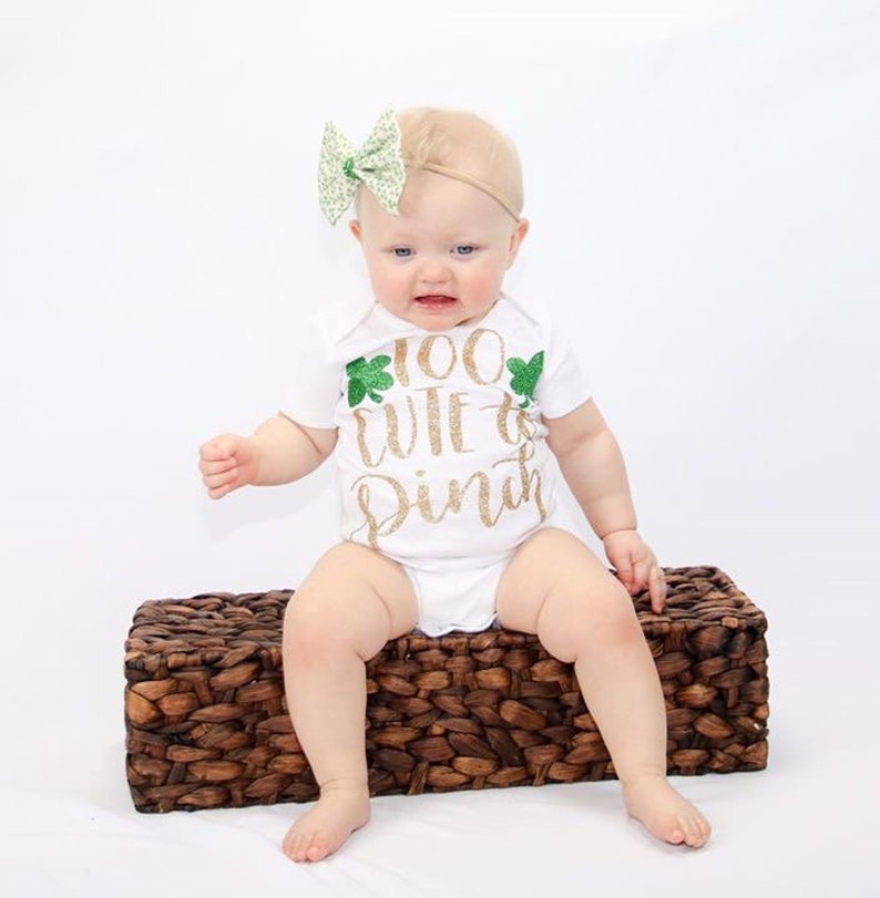 May include: A white baby onesie with a gold glitter design that says "Too Cute to Pinch" with green shamrock accents. The baby is wearing a green and white bow headband.