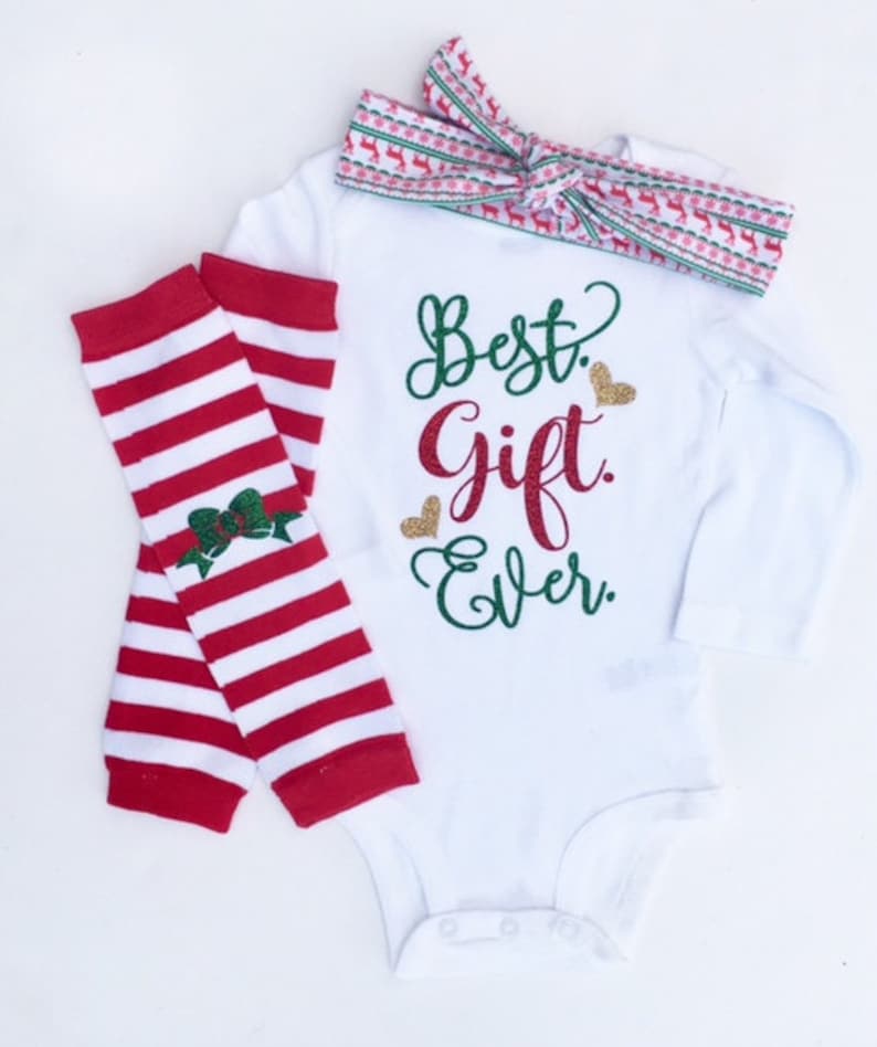 Puede incluir: Un mono blanco de manga larga con el texto "Best Gift Ever" en purpurina verde y dorada. El mono se combina con polainas a rayas rojas y blancas y una diadema roja y blanca con estampado de reno.
