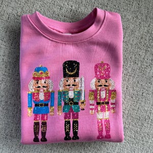 May include: Pink sweatshirt with a glittery design of three nutcracker soldiers. The nutcrackers are wearing different colored uniforms and hats.