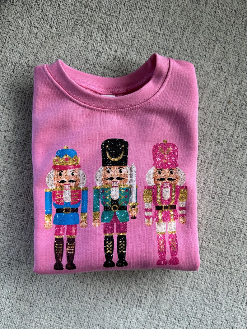 May include: Pink sweatshirt with a glittery design of three nutcracker soldiers. The nutcrackers are wearing different colored uniforms and hats.