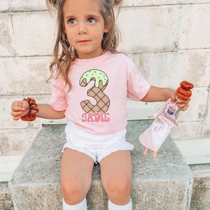 May include: A young girl wearing a pink t-shirt with a green and brown ice cream cone number "3" and the name "SADIE" printed on it. She is holding a brown hair scrunchie in her hand and a doll in the other.