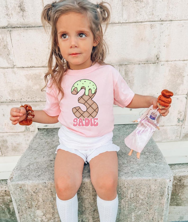 May include: A young girl wearing a pink t-shirt with a green and brown ice cream cone number "3" and the name "SADIE" printed on it. She is holding a brown hair scrunchie in her hand and a doll in the other.
