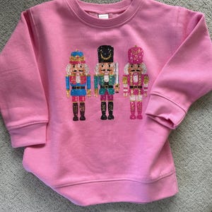 May include: Pink sweatshirt with a festive design of three nutcracker soldiers in glitter. The nutcrackers are wearing traditional costumes with gold accents.