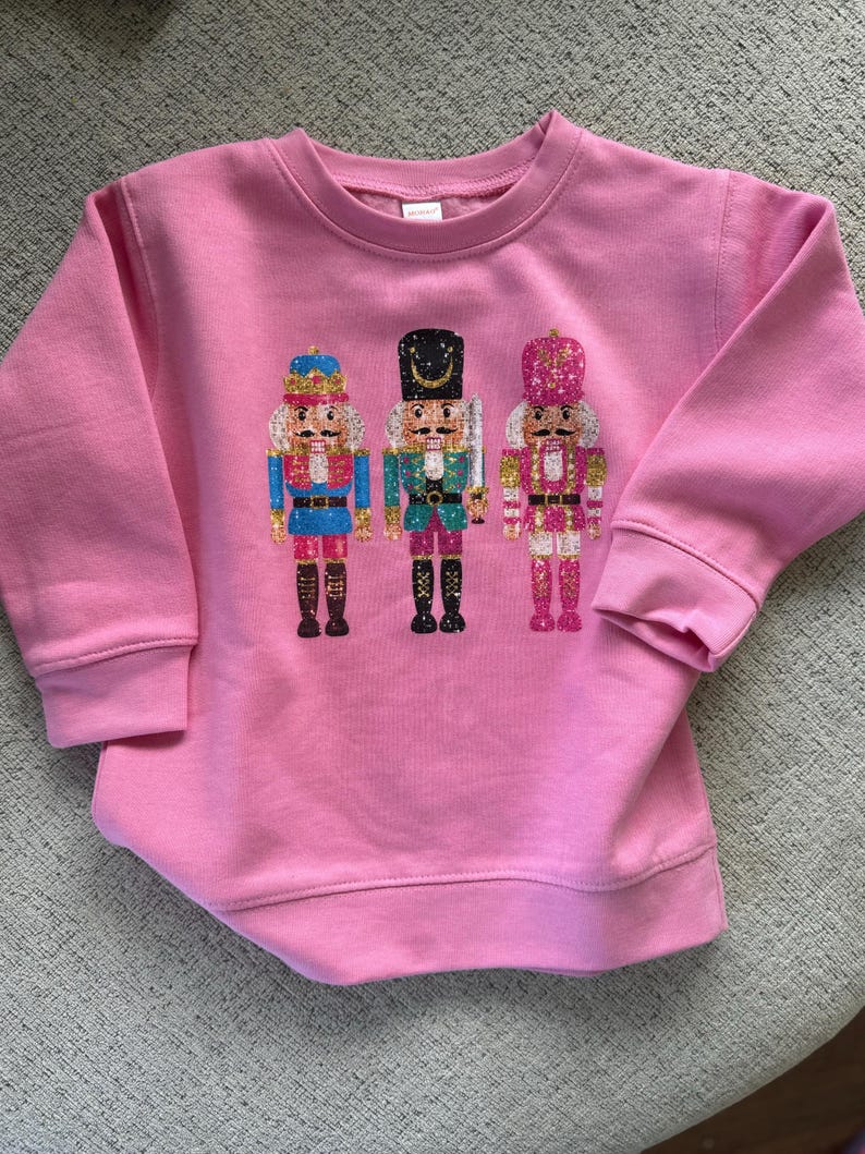 May include: Pink sweatshirt with a festive design of three nutcracker soldiers in glitter. The nutcrackers are wearing traditional costumes with gold accents.