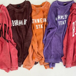 May include: Five long-sleeved shirts with different college logos. The shirts are in various colors, including red, brown, orange, purple, and pink. The logos are white and include the words "U of A", "BEAVER", "NORTHWESTERN STATE", "SOUTHWESTERN STATE", and "RICE".