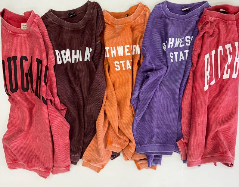 May include: Five long-sleeved shirts with different college logos. The shirts are in various colors, including red, brown, orange, purple, and pink. The logos are white and include the words "U of A", "BEAVER", "NORTHWESTERN STATE", "SOUTHWESTERN STATE", and "RICE".