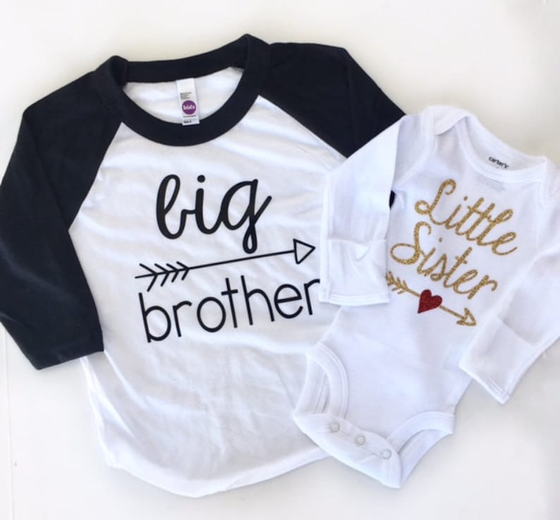 Puede incluir: Dos camisas blancas con letras negras y doradas. La camisa m&aacute;s grande dice "hermano mayor" con una flecha apuntando hacia la derecha. La camisa m&aacute;s peque&ntilde;a dice "Hermana peque&ntilde;a" con una flecha apuntando hacia la derecha y un coraz&oacute;n rojo.