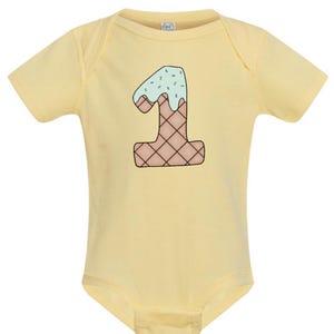 May include: A yellow baby onesie with a graphic of the number one shaped like an ice cream cone with green icing and sprinkles.