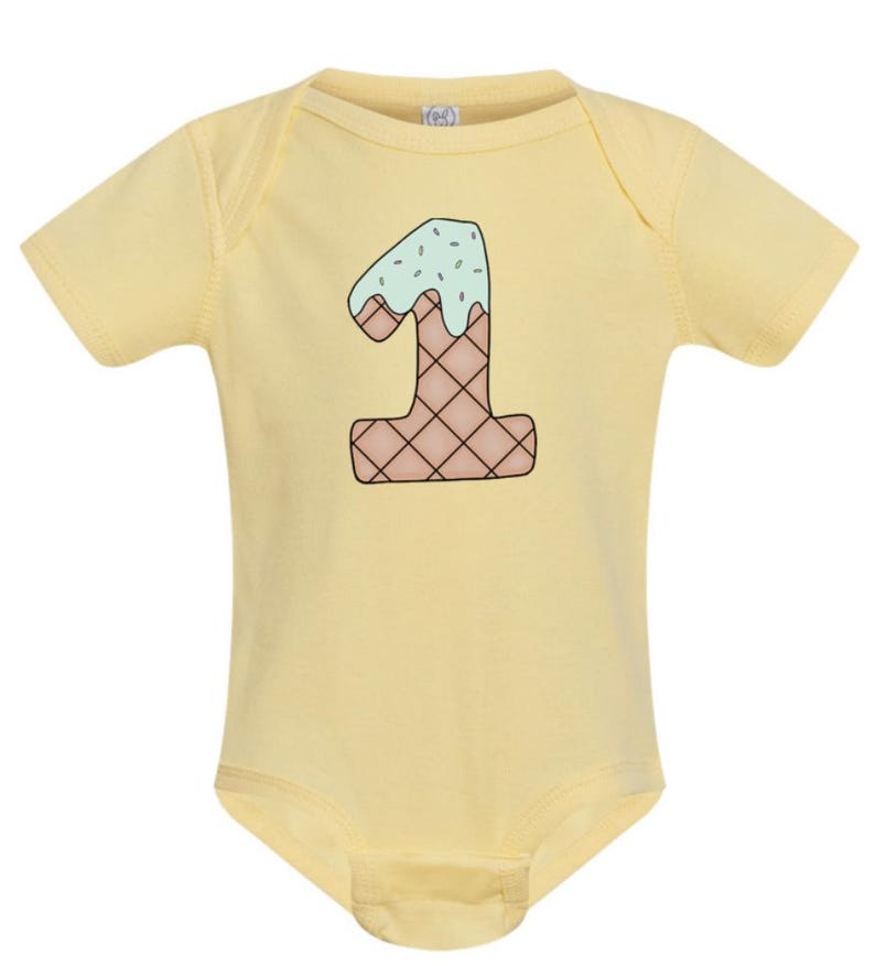 May include: A yellow baby onesie with a graphic of the number one shaped like an ice cream cone with green icing and sprinkles.