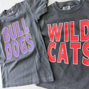 K&ouml;nnte beinhalten: Zwei graue T-Shirts mit unterschiedlich farbigem Text. Das erste Shirt sagt "Bulldogs" in Lila mit gelber Umrandung. Das zweite Shirt sagt "Wildcats" in Rot mit wei&szlig;er Umrandung.