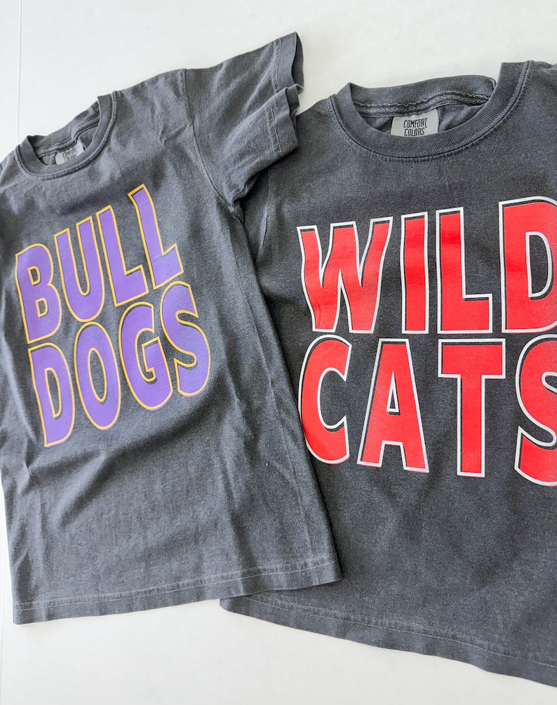 K&ouml;nnte beinhalten: Zwei graue T-Shirts mit unterschiedlich farbigem Text. Das erste Shirt sagt "Bulldogs" in Lila mit gelber Umrandung. Das zweite Shirt sagt "Wildcats" in Rot mit wei&szlig;er Umrandung.