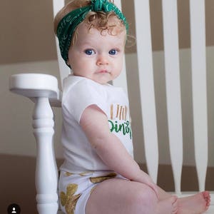 Puede incluir: Una ni&ntilde;a peque&ntilde;a con un body blanco con letras doradas brillantes que dicen "Little Irish" y pantalones cortos con estampado de tr&eacute;bol verde. Lleva una diadema verde con letras blancas y tiene el pelo rubio rizado. Est&aacute; sentada en una silla blanca.