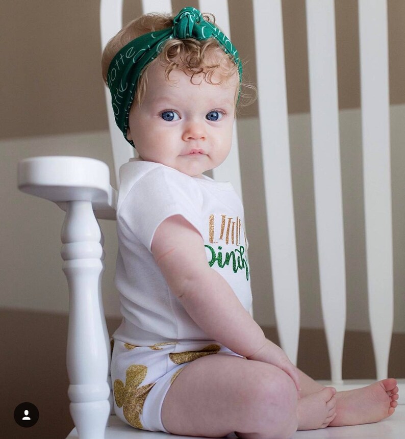 Puede incluir: Una ni&ntilde;a peque&ntilde;a con un body blanco con letras doradas brillantes que dicen "Little Irish" y pantalones cortos con estampado de tr&eacute;bol verde. Lleva una diadema verde con letras blancas y tiene el pelo rubio rizado. Est&aacute; sentada en una silla blanca.