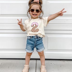 May include: A young child wearing a light beige t-shirt with a pink and brown ice cream cone shaped number "2" on the front. The child is wearing blue denim shorts and light beige sneakers. The child is smiling and giving a peace sign with both hands.
