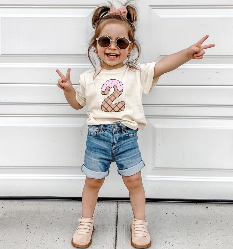 May include: A young child wearing a light beige t-shirt with a pink and brown ice cream cone shaped number "2" on the front. The child is wearing blue denim shorts and light beige sneakers. The child is smiling and giving a peace sign with both hands.