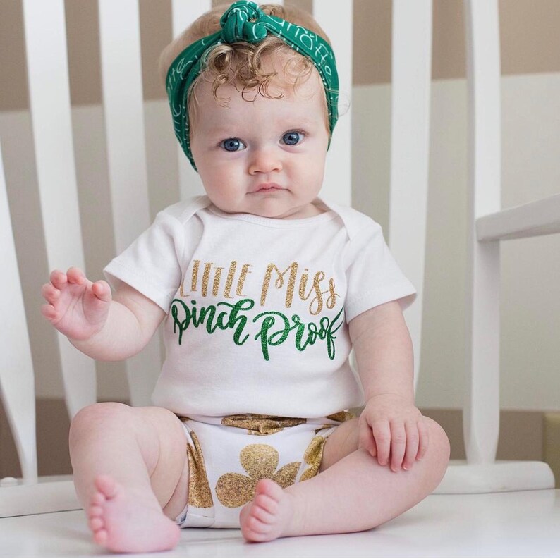 Puede incluir: Una ni&ntilde;a peque&ntilde;a con un body blanco con el texto "Little Miss Pinch Proof" en purpurina verde y dorada. Lleva una diadema verde y tiene el pelo rubio rizado.