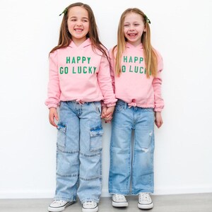 Puede incluir: Dos chicas con sudaderas rosas con el texto "Happy Go Lucky" en letras verdes. Llevan pantalones vaqueros azules y zapatillas blancas.