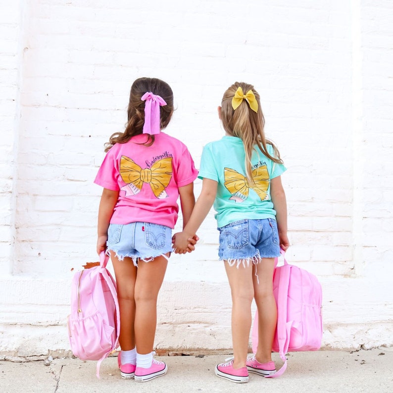 May include: Two girls wearing pink and turquoise t-shirts with a yellow bow graphic on the back. The shirts say "Kindergarten" and "First Grade". They are wearing denim shorts and carrying pink backpacks.