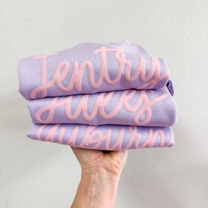 以下が含まれることがあります： ピンクの文字で書かれたラベンダー色のTシャツが3枚重ねられています。Tシャツは折り畳まれており、文字は「Jentry」、「Hayes」、「Auburn」と書いてあります。
