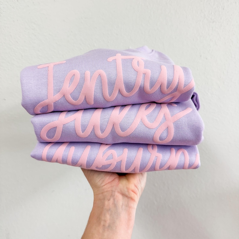 以下が含まれることがあります： ピンクの文字で書かれたラベンダー色のTシャツが3枚重ねられています。Tシャツは折り畳まれており、文字は「Jentry」、「Hayes」、「Auburn」と書いてあります。