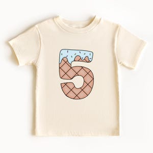 May include: A cream colored t-shirt with a cartoon ice cream cone shaped like the number 5. The ice cream is blue with pink sprinkles.