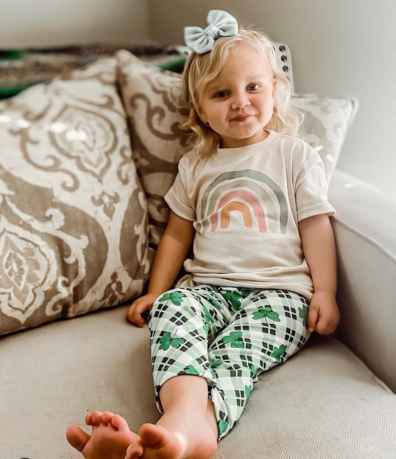 Op de afbeelding: Een jong kind draagt een lichtbeige t-shirt met een regenboog-print en een groen-wit geruite broek met klavertjesvier-ontwerpen. Het kind zit op een bank met een lichtbruin, gepatroneerd kussen.