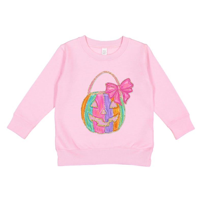 Puede incluir: Una sudadera rosa de manga larga con una bolsa de truco o trato en forma de calabaza colorida con un lazo rosa en la parte superior.