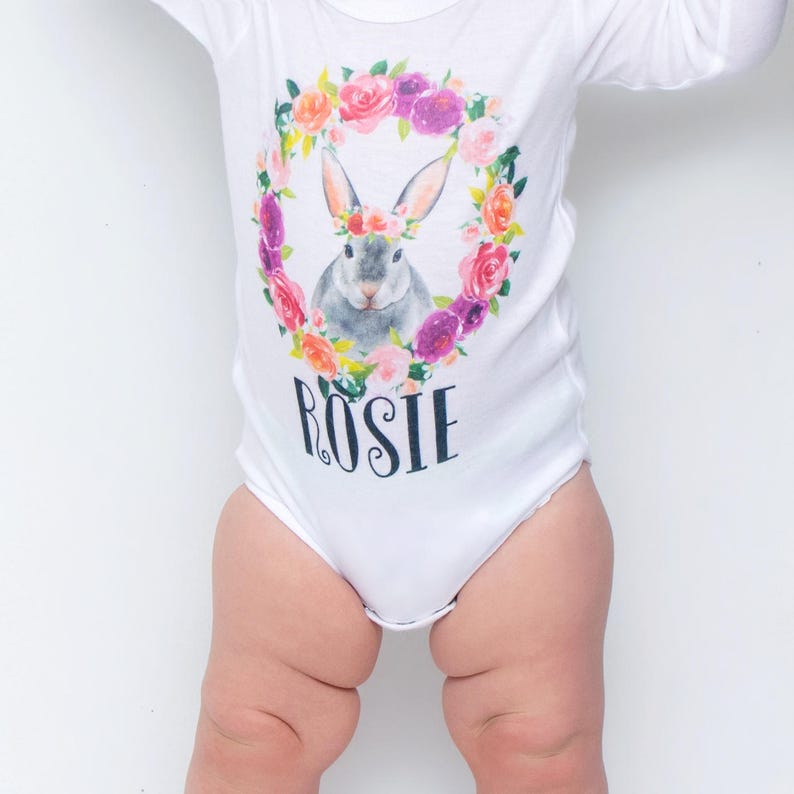 May include: White long-sleeved bodysuit with a floral wreath and a bunny wearing a flower crown. The text "ROSIE" is printed below the wreath.