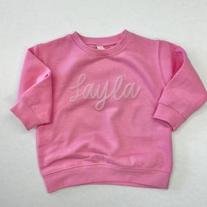 以下が含まれることがあります： ピンクのトレーナーに、白い立体文字で「Layla」と書いてあります。