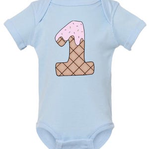 May include: Light blue baby bodysuit with a number one shaped like an ice cream cone with pink frosting and sprinkles.