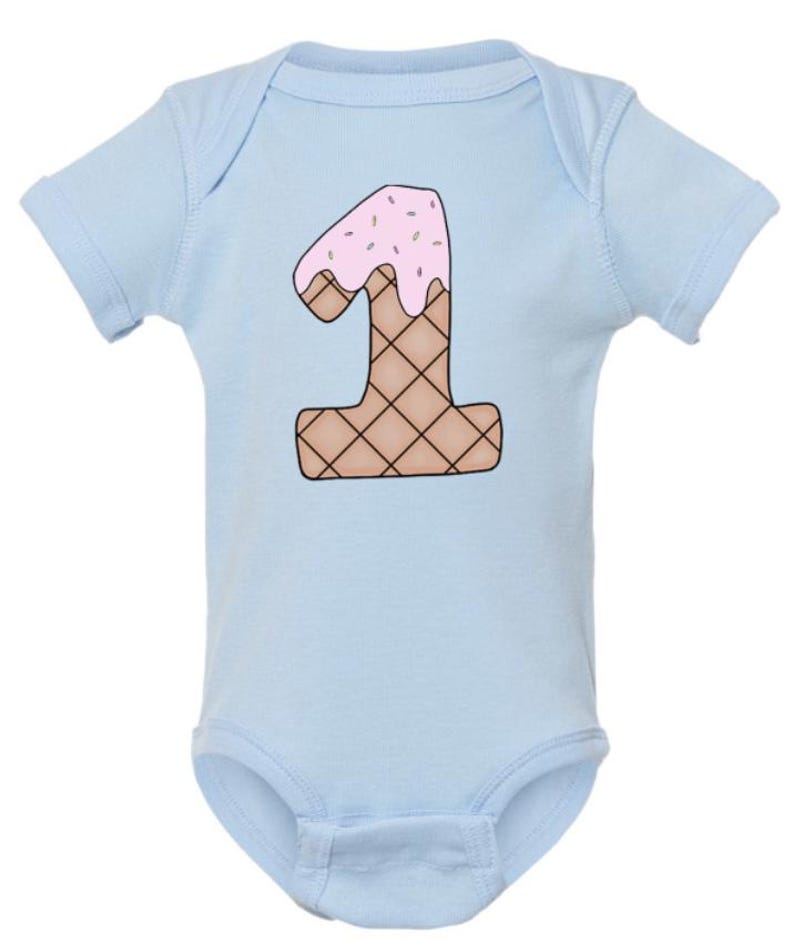 May include: Light blue baby bodysuit with a number one shaped like an ice cream cone with pink frosting and sprinkles.