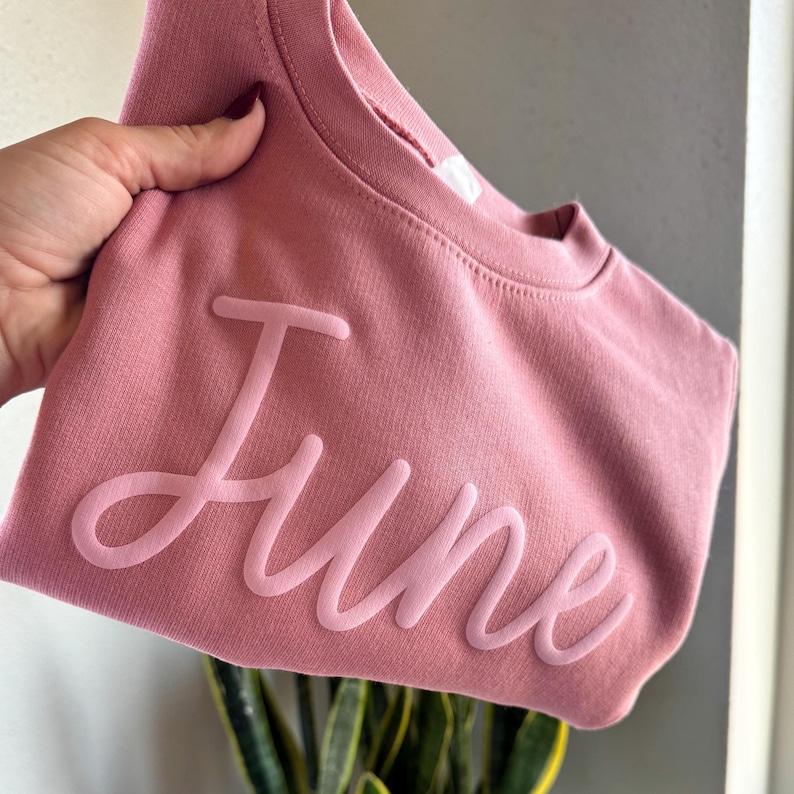 以下が含まれることがあります： 白い立体文字で「June」と書かれたピンク色のスウェットシャツです。