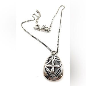 May include: Silver-toned necklace with a teardrop pendant. The pendant features a four-petal design with a central clear stone and is surrounded by a dotted border. The chain is a delicate, woven style.