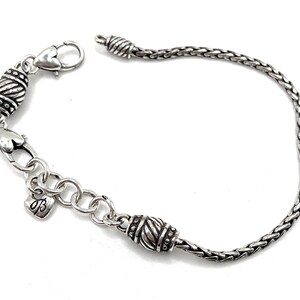 May include: A silver bracelet with a braided chain and two decorative silver beads. The bracelet has a heart-shaped clasp and a small charm with the letter "B" on it.