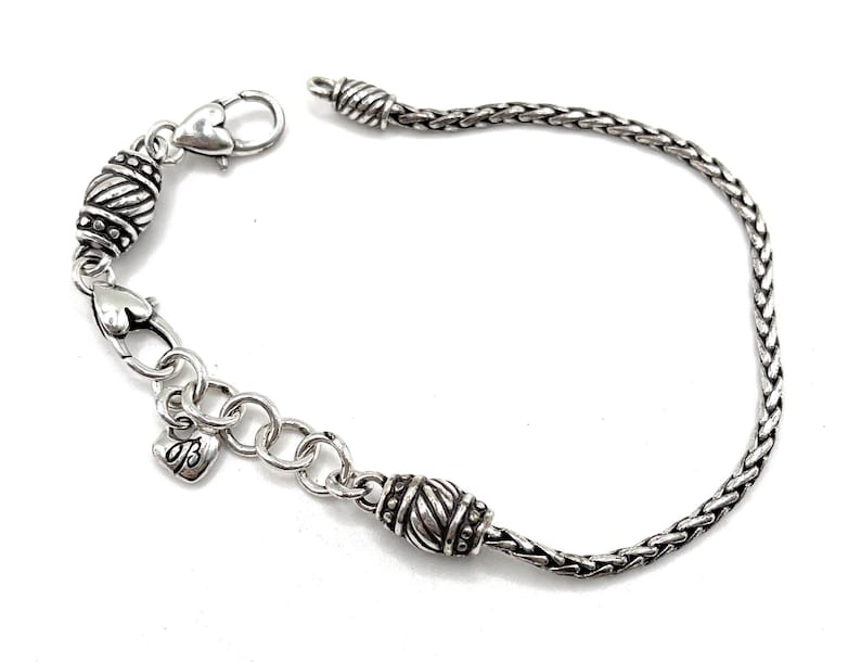 May include: A silver bracelet with a braided chain and two decorative silver beads. The bracelet has a heart-shaped clasp and a small charm with the letter "B" on it.