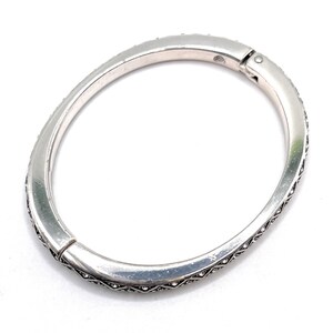 May include: Silver-toned hinged bangle bracelet. The bracelet is adorned with small, clear crystals along the outer edges. The clasp is visible at the top, and the bracelet has a slightly oval shape.