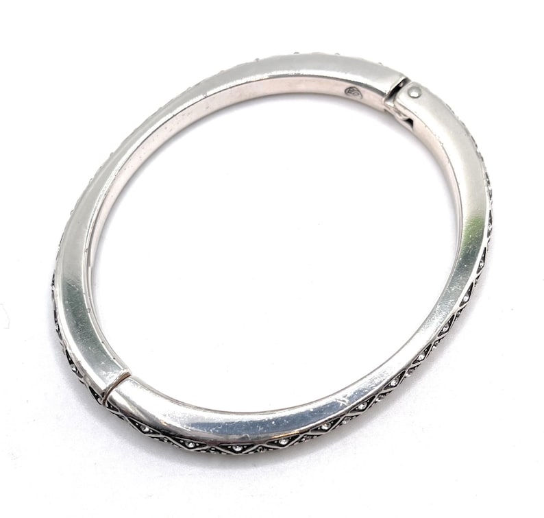 May include: Silver-toned hinged bangle bracelet. The bracelet is adorned with small, clear crystals along the outer edges. The clasp is visible at the top, and the bracelet has a slightly oval shape.