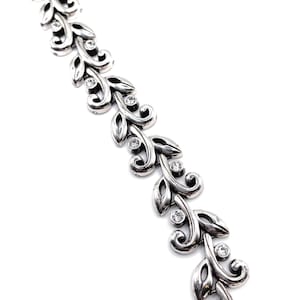 Puede incluir: Una pulsera plateada con un dise&ntilde;o de hojas y gemas transparentes. La pulsera presenta una serie de hojas estilizadas conectadas por eslabones curvos, con una gema redonda en el centro de cada hoja. La pulsera se muestra sobre un fondo blanco.