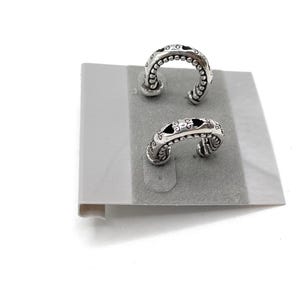 May include: Two silver-toned hoop earrings with intricate designs. Each earring features a curved shape with small, raised beads along the outer edge and decorative cutouts. The earrings are displayed on a gray, felt-like surface.
