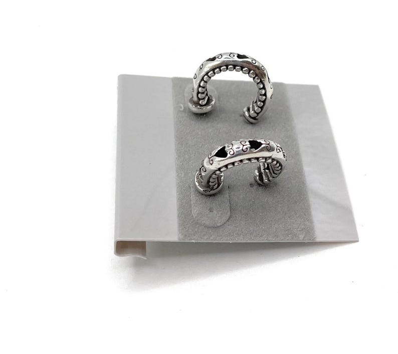 May include: Two silver-toned hoop earrings with intricate designs. Each earring features a curved shape with small, raised beads along the outer edge and decorative cutouts. The earrings are displayed on a gray, felt-like surface.