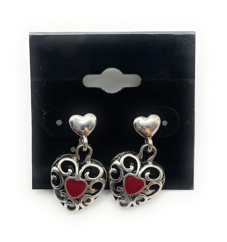 May include: Silver-coloured heart-shaped earrings with a red heart centre. The earrings feature a filigree design and are displayed on a black earring card.