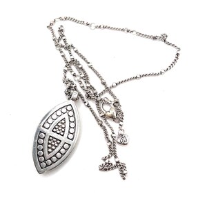 Puede incluir: Un collar plateado con un colgante ovalado grande. El colgante presenta un dise&ntilde;o geom&eacute;trico con c&iacute;rculos y un patr&oacute;n central. El collar tiene m&uacute;ltiples estilos de cadena, incluyendo un dije en forma de coraz&oacute;n y un peque&ntilde;o dije decorativo.