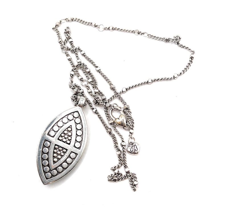 Puede incluir: Un collar plateado con un colgante ovalado grande. El colgante presenta un dise&ntilde;o geom&eacute;trico con c&iacute;rculos y un patr&oacute;n central. El collar tiene m&uacute;ltiples estilos de cadena, incluyendo un dije en forma de coraz&oacute;n y un peque&ntilde;o dije decorativo.