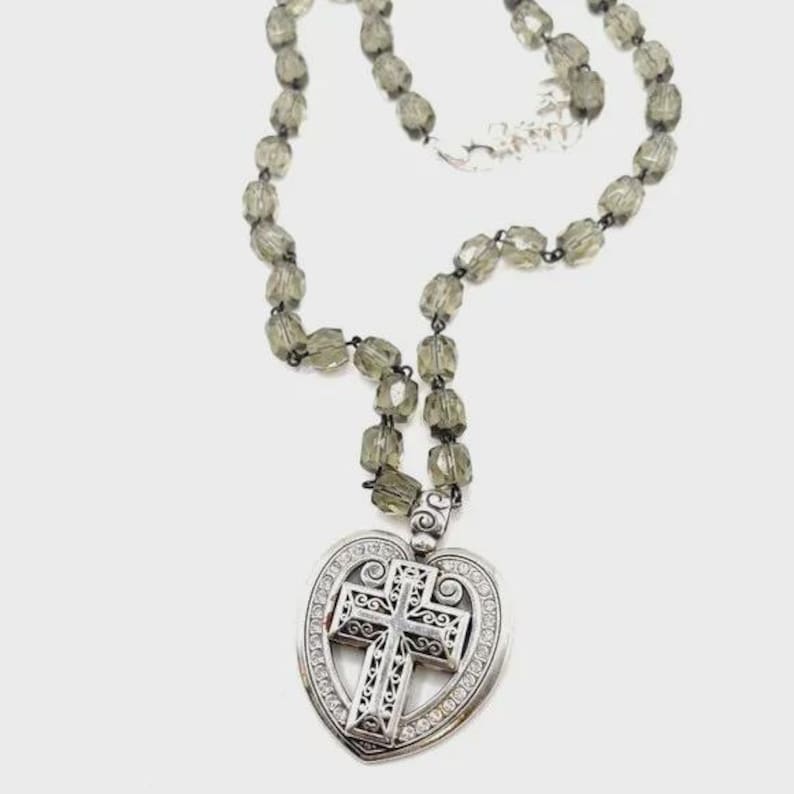 Peut inclure: Un collier argent&eacute; avec un pendentif en forme de c&oelig;ur et un motif de croix. Le pendentif est orn&eacute; de cristaux transparents. Le collier a une cha&icirc;ne perl&eacute;e avec des perles facett&eacute;es vert olive.